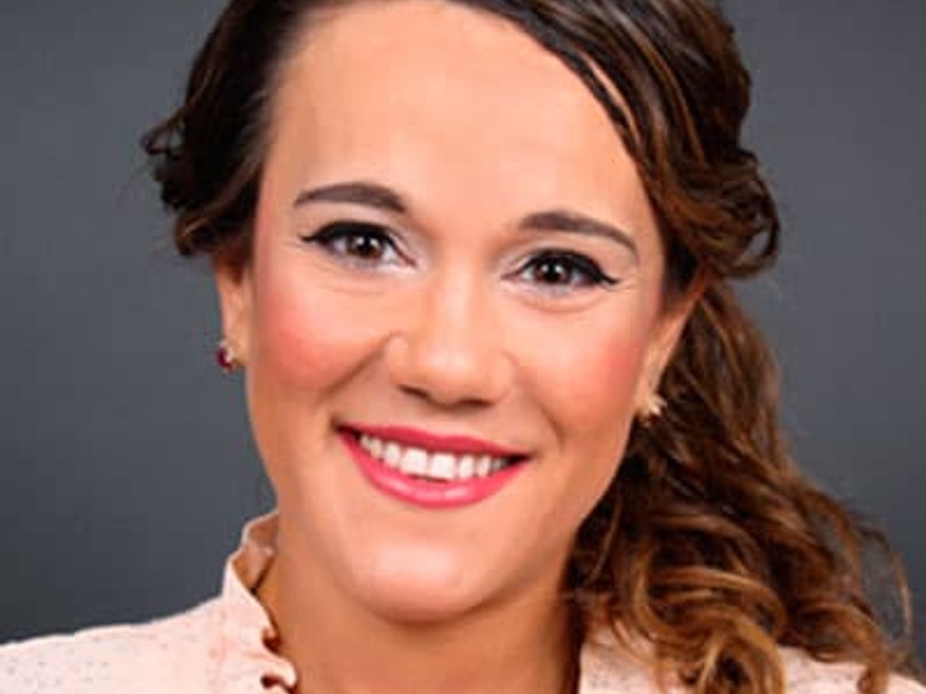 A portrait of a smiling woman with curly hair, wearing a light pink blouse and subtle makeup.