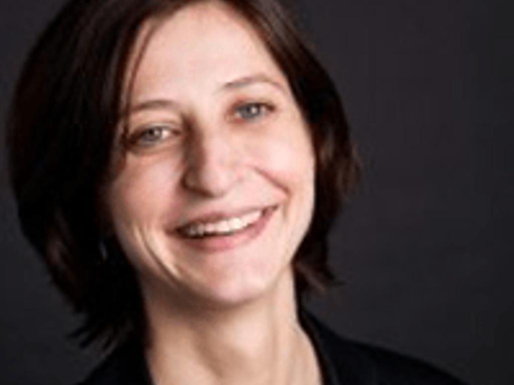 Portrait of a smiling woman with short hair, wearing a black blazer against a dark background.