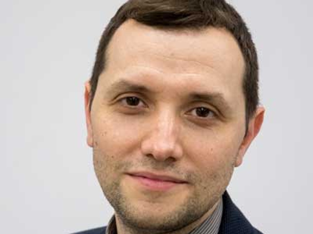 A portrait of a man with a slight smile, wearing a dark jacket over a checked shirt.