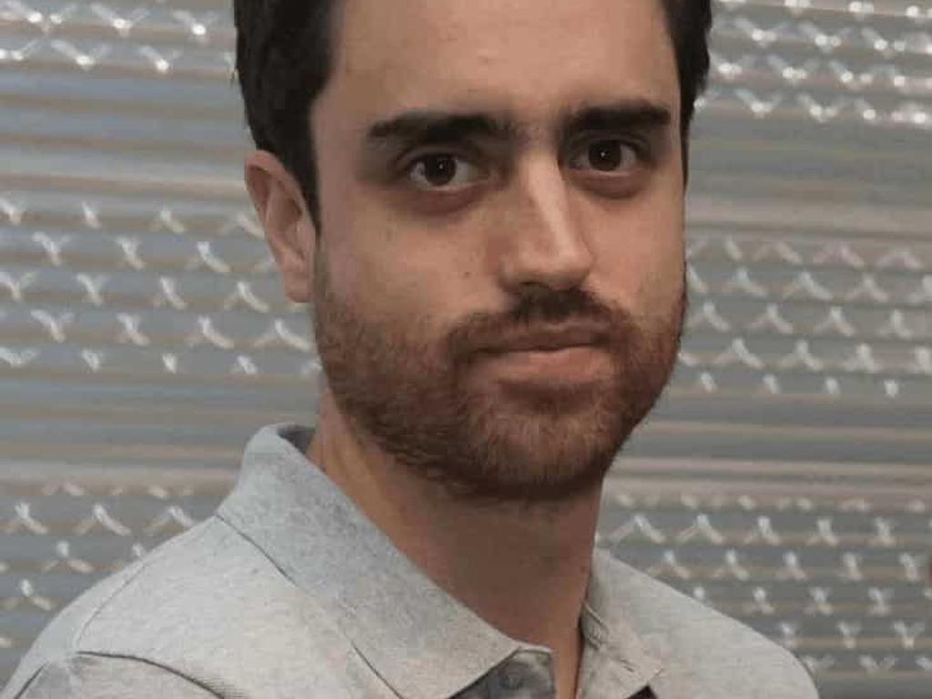 A man with a beard and short dark hair, wearing a grey shirt, standing indoors with a patterned backdrop.