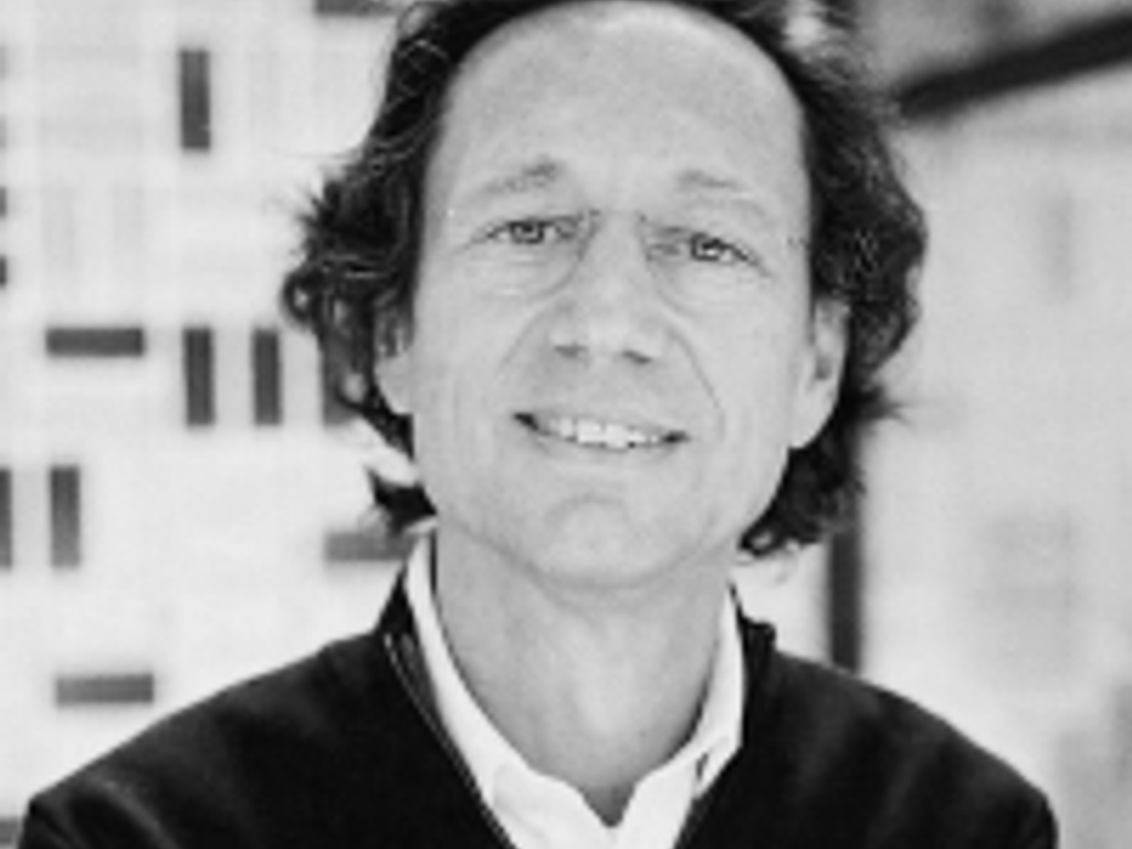 Black and white photo of a smiling middle-aged man with long hair, standing indoors with blurred buildings in the background.