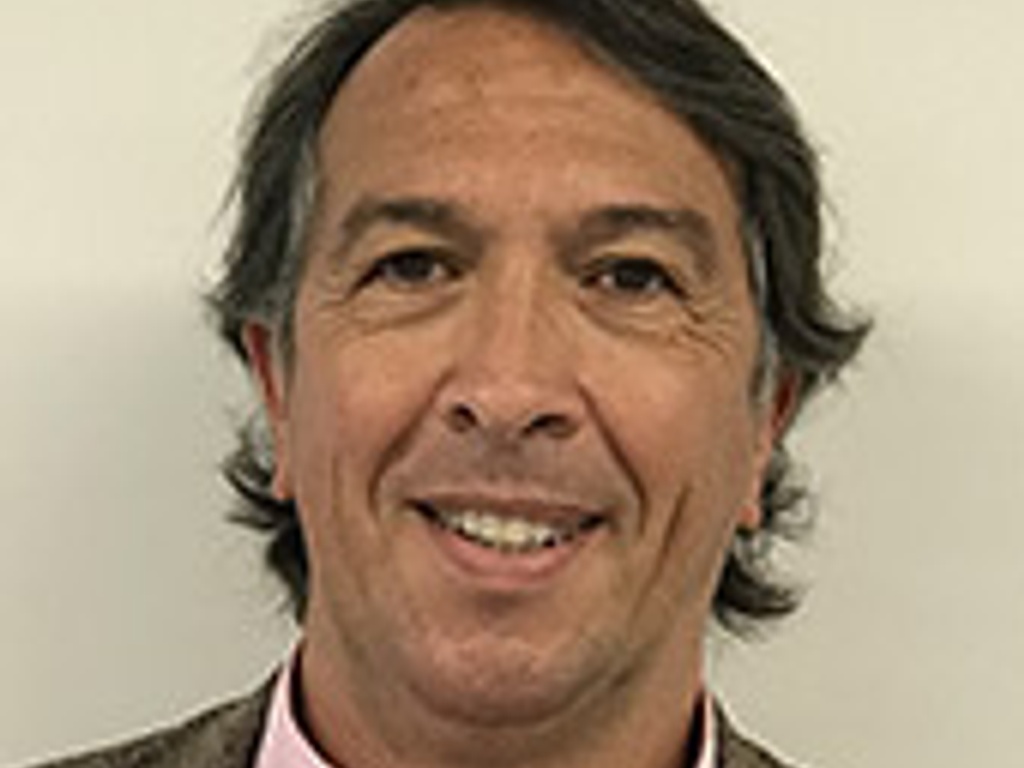 A portrait of a smiling middle-aged man with long hair wearing a pink shirt and a tweed jacket.