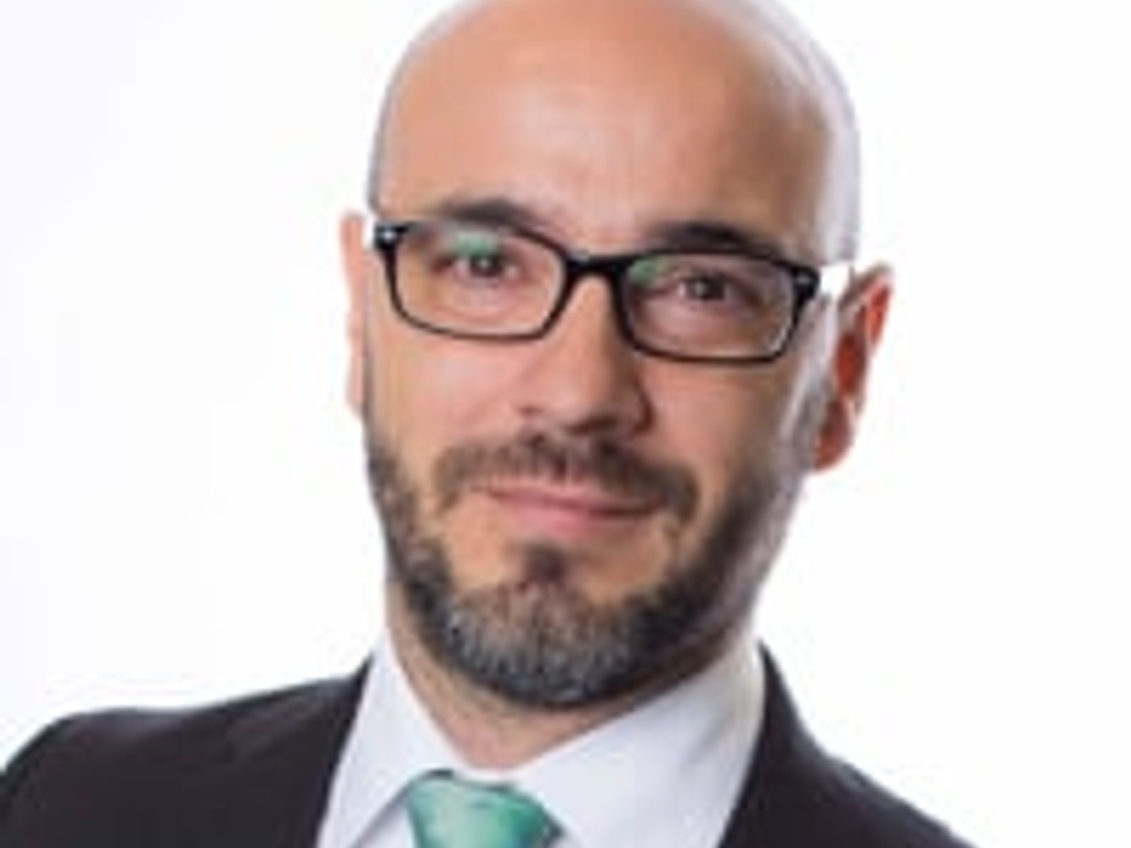 A professional portrait of a bald man with a beard, wearing a black suit and a green tie.