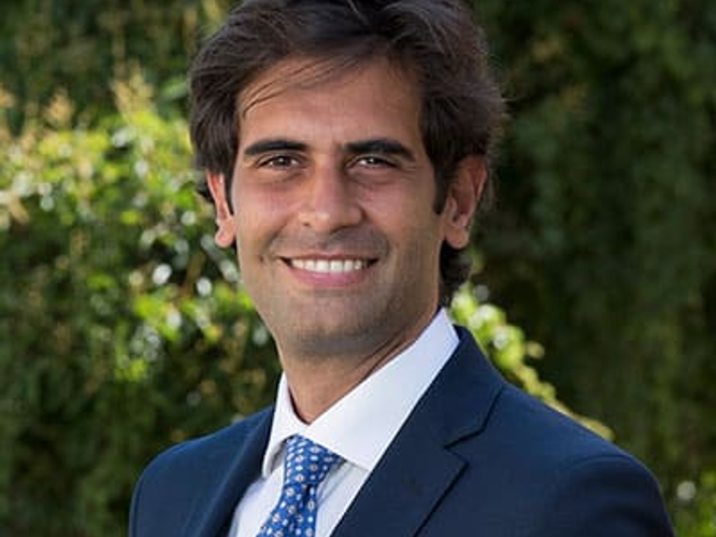 A smiling man with brown hair, wearing a blue suit and blue polka dot tie, standing outdoors with greenery in the background.