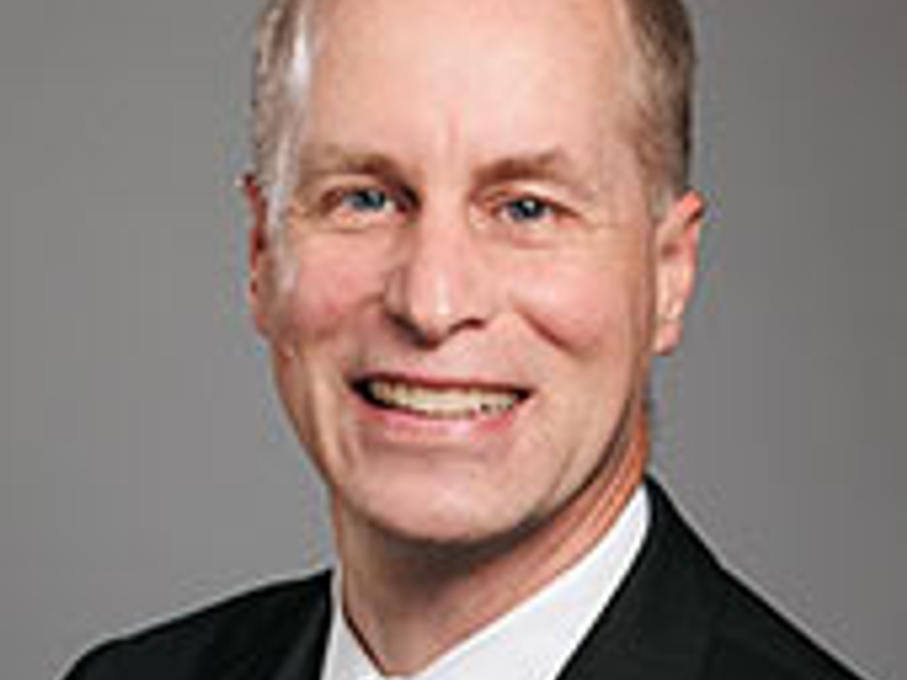 A professional headshot of a smiling bald man wearing a black suit and a blue striped tie.