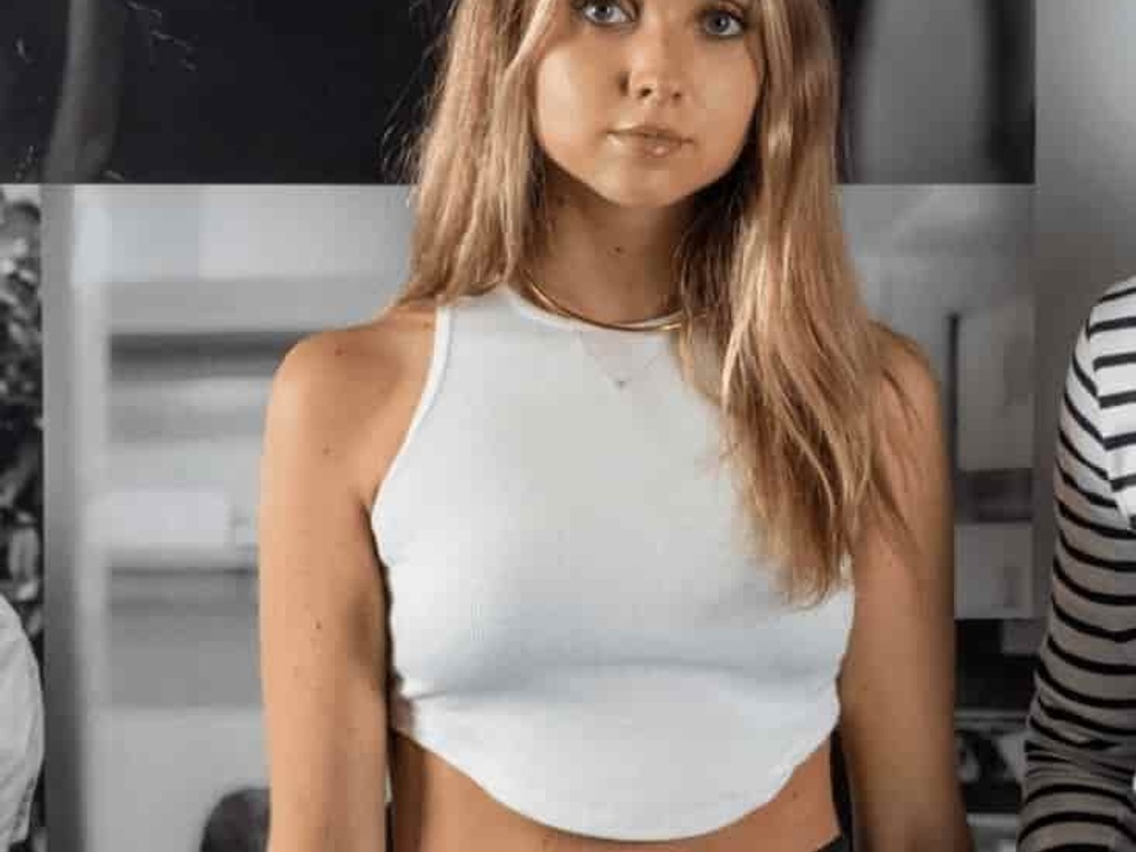 A young woman with light brown hair wearing a white sleeveless top and black pants, standing against a black and white photo backdrop.