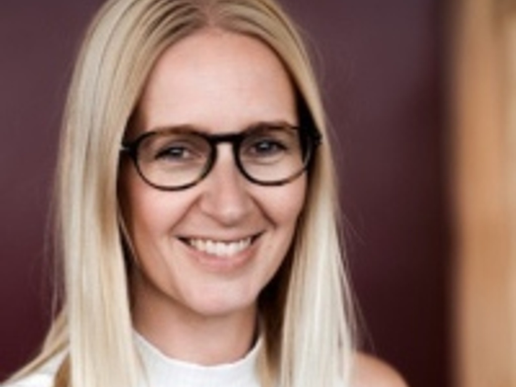 A smiling woman with blonde hair wearing glasses and a white sleeveless top.