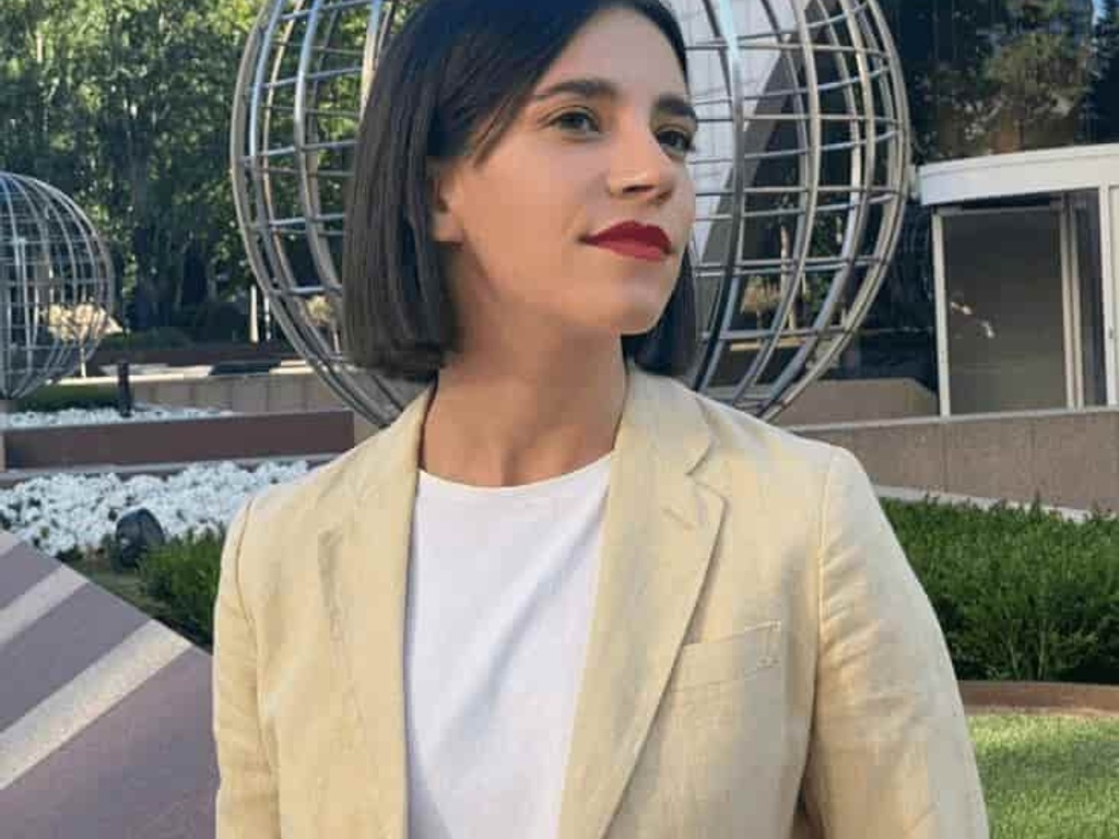 A woman standing outdoors in a business district, dressed in a cream blazer, looking towards her left with a thoughtful expression.