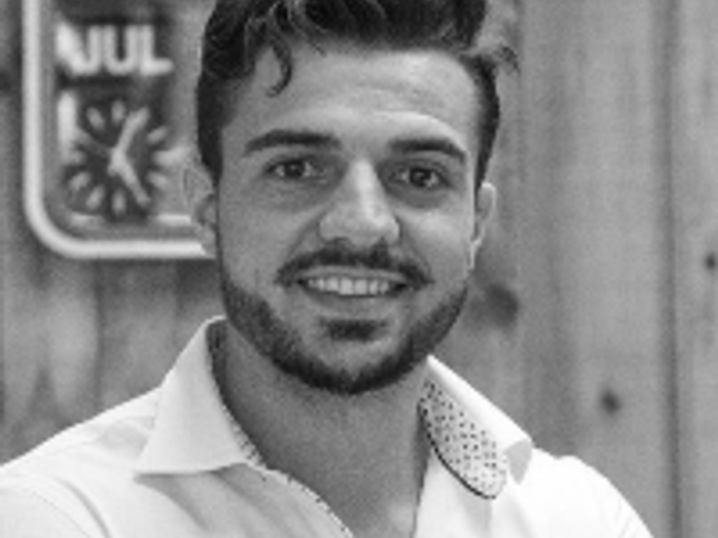 A black and white photo of a smiling young man with styled hair, wearing a white shirt.