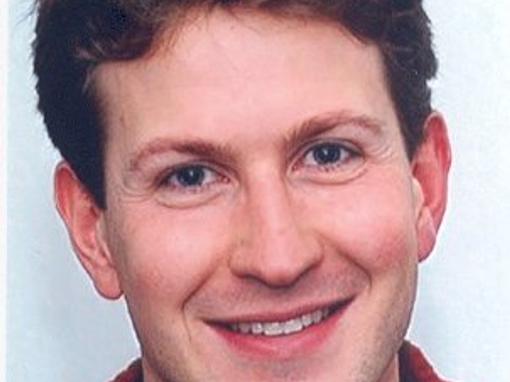 A close-up portrait of a smiling man with short brown hair, wearing a red jacket.