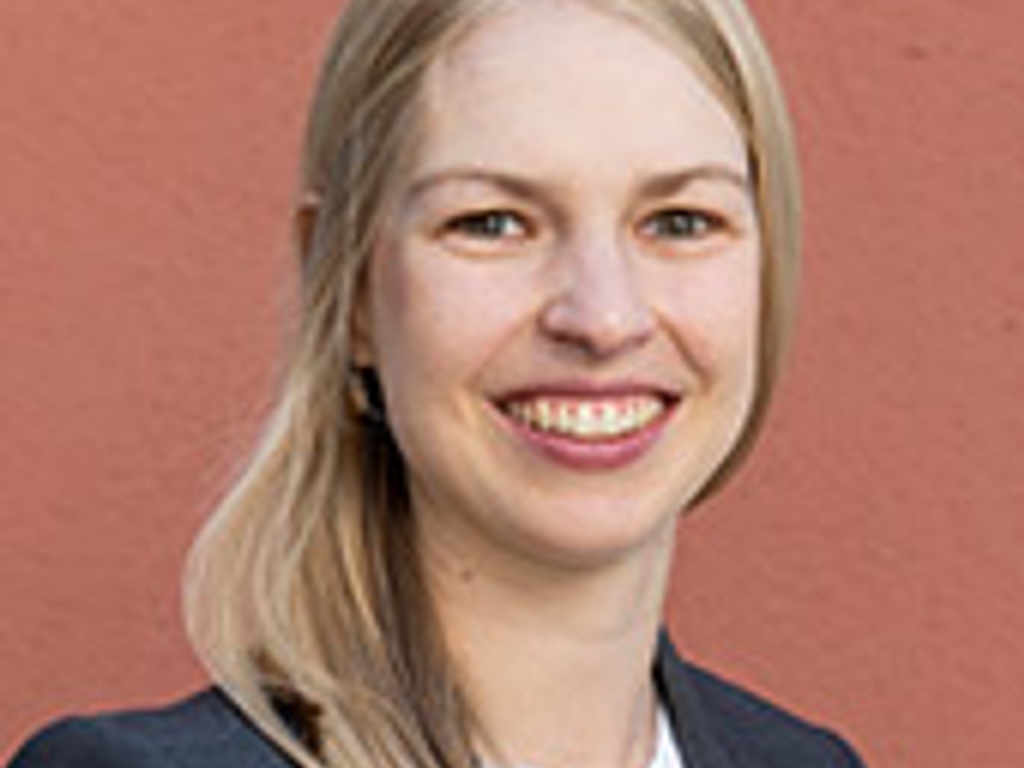 A portrait of a smiling young woman with blond hair, wearing a dark blazer over a light blouse, standing against a red background.