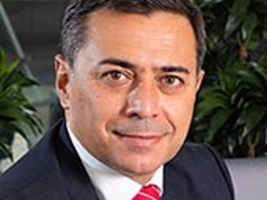 A professional portrait of a man wearing a suit and a red tie, smiling in an office setting with plants in the background.