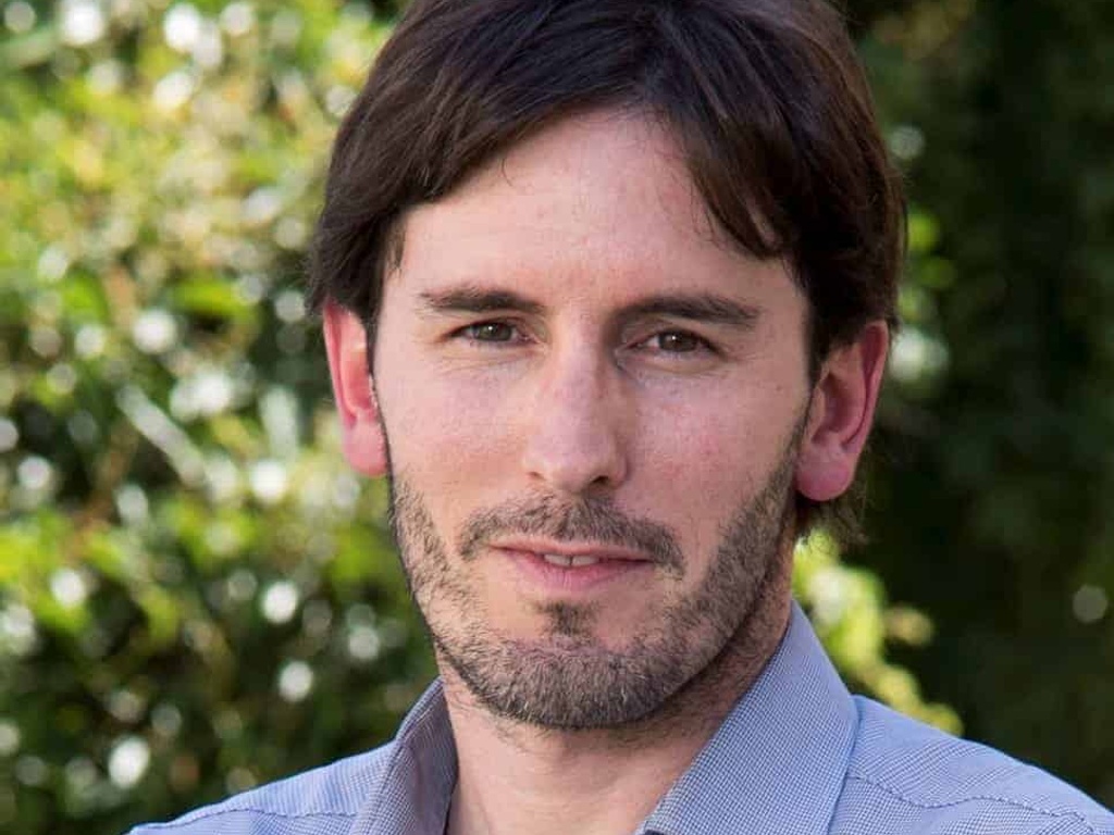 A portrait of a man with a beard in a blue shirt standing outdoors with trees in the background.