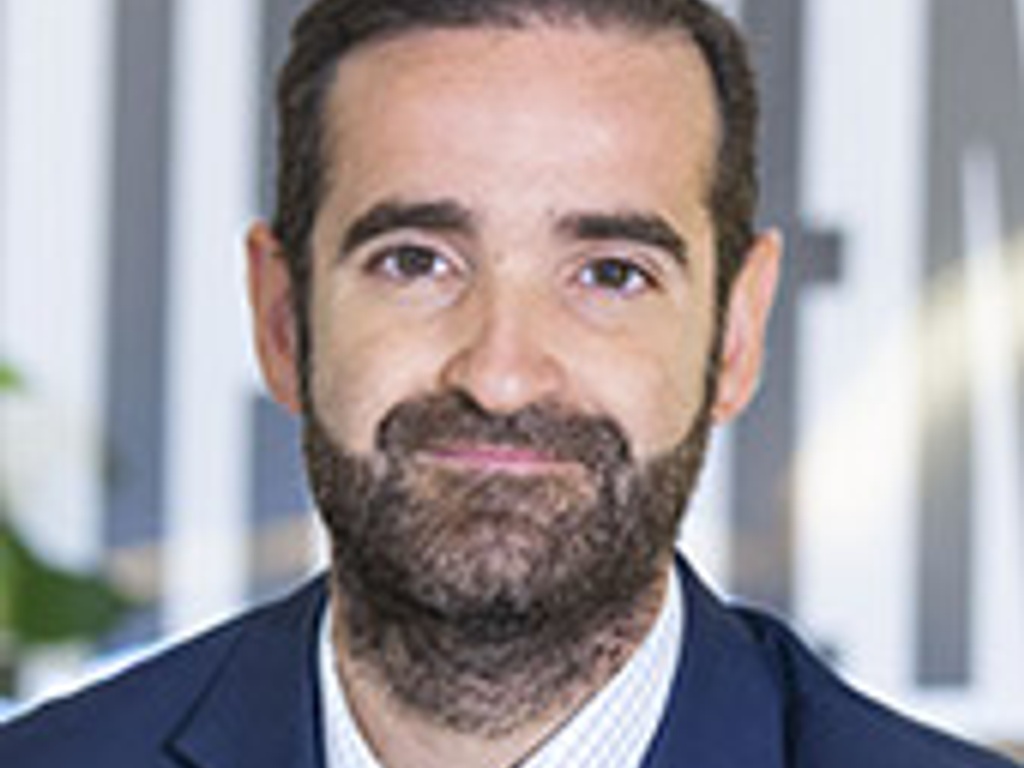 Portrait of a man with a beard, wearing a suit and checkered shirt, smiling in an office setting.