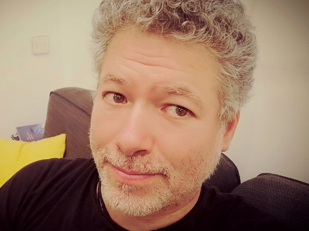 A close-up portrait of a middle-aged man with curly gray hair and a slight smile, sitting in a room with a yellow pillow and blue sofa in the background.