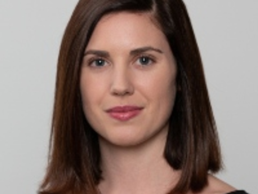 A professional headshot of a woman with shoulder-length brown hair, wearing a black top and minimal makeup, looking directly at the camera.