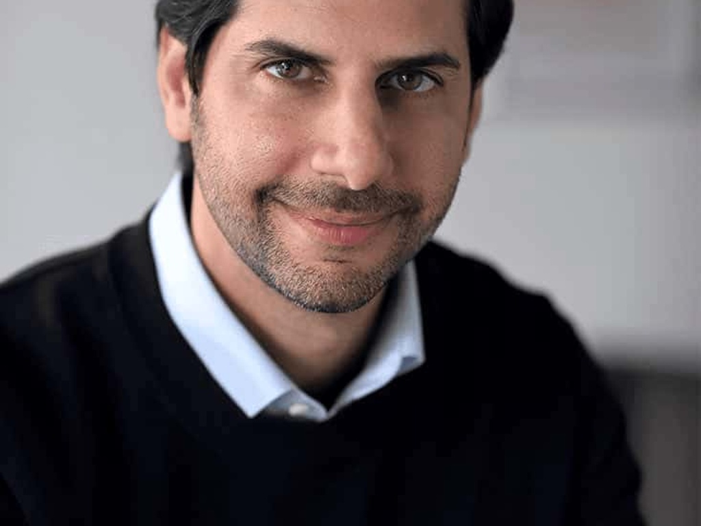 A portrait of a smiling man with dark hair, wearing a dark sweater and a white shirt.