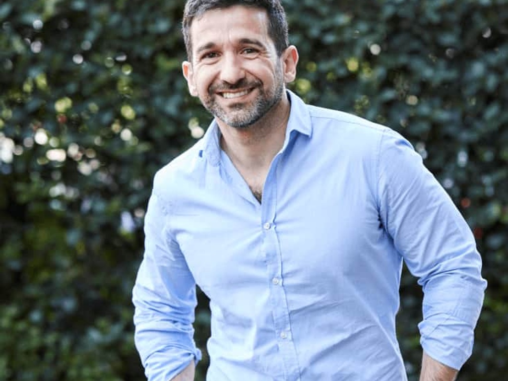 A smiling man standing outdoors wearing a blue shirt.