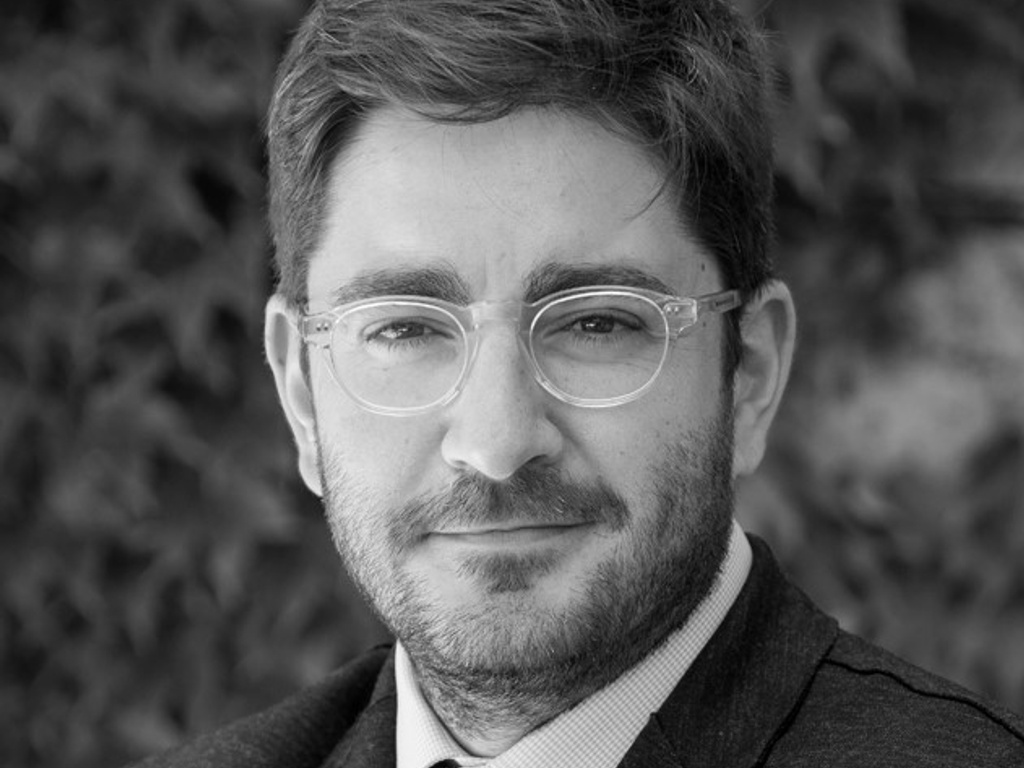 A black and white portrait of a man with glasses, wearing a suit and a polka dot tie, standing in front of a leafy background.