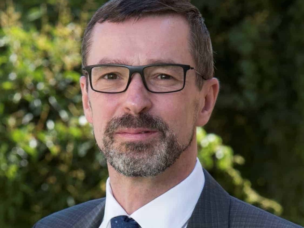 Portrait of a smiling middle-aged man wearing glasses and a suit with a tie, standing outdoors.