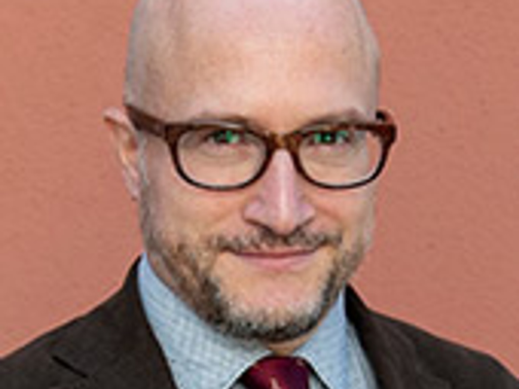 Portrait of a bald man wearing glasses and a suit with a burgundy tie standing against a pink background.