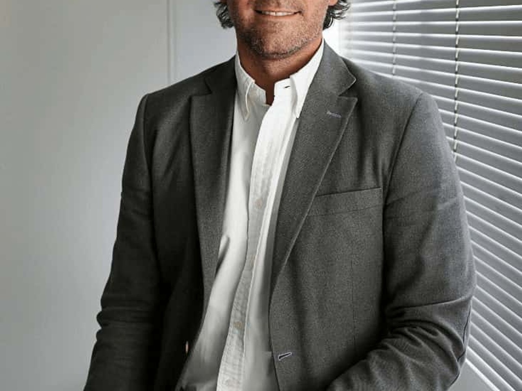 A man in a gray blazer and white shirt sits by a window with partially closed blinds, smiling subtly towards the camera.