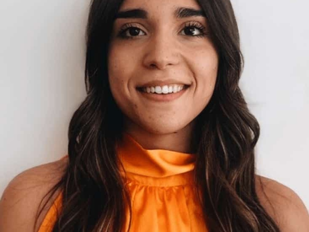 A portrait of a smiling woman with long brown hair, wearing an orange top.