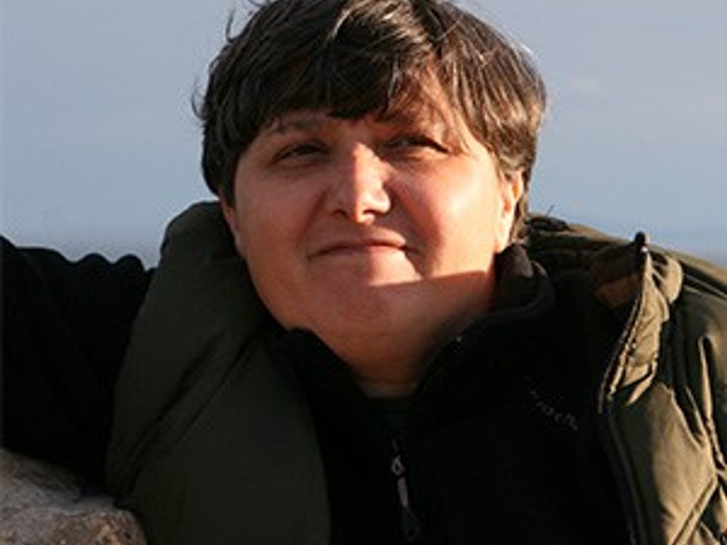 A smiling person with short hair wearing a dark jacket, resting outdoors with a bright backdrop.