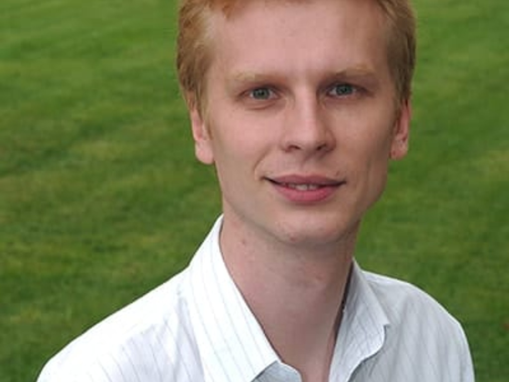 Portrait of a smiling young man with short red hair, wearing a white striped shirt, standing outdoors.