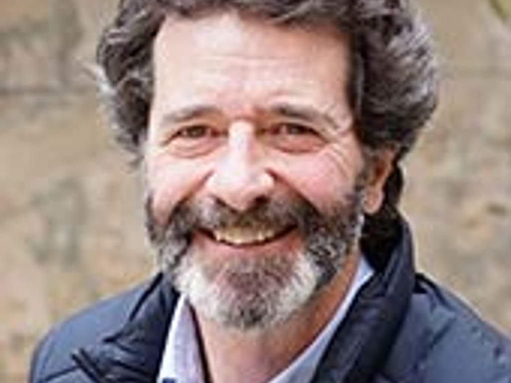 A smiling older man with a beard and curly hair wearing a blue jacket over a buttoned shirt