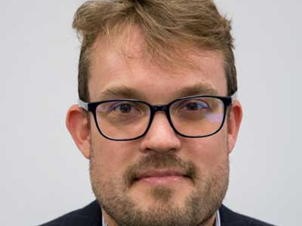 Portrait of a man with glasses and stubble, wearing a dark blazer.