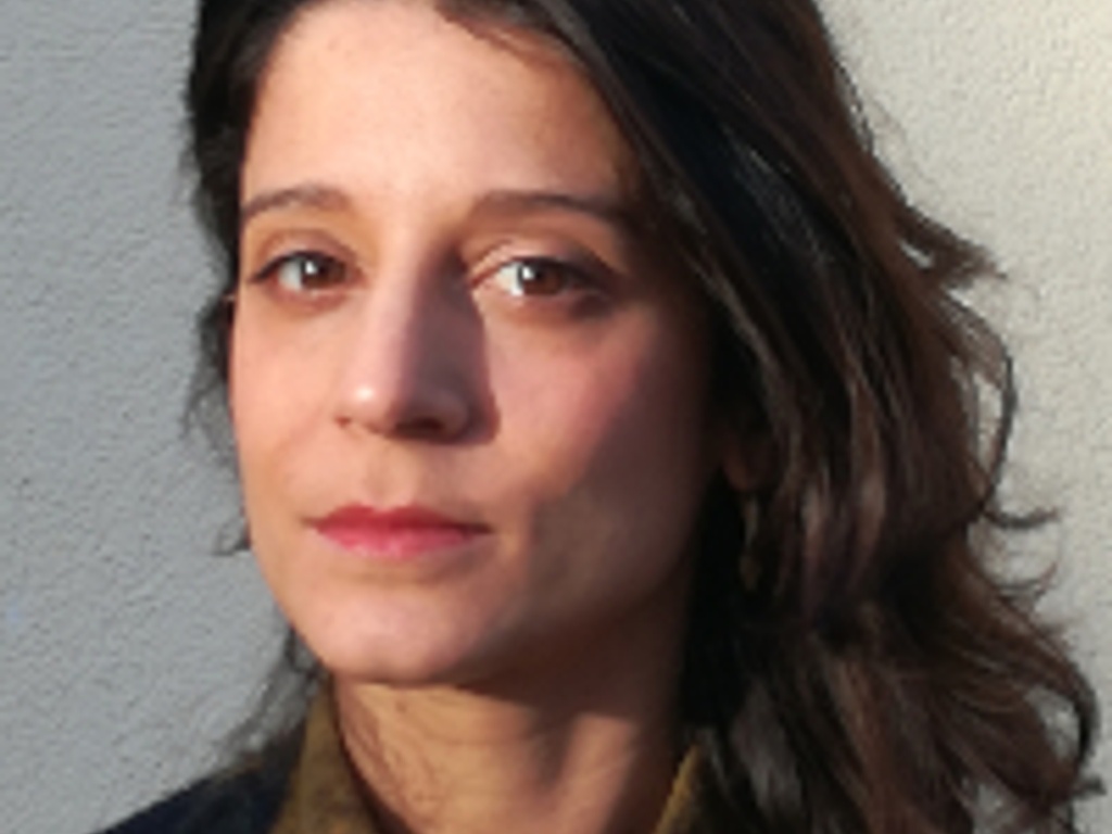 A portrait of a woman with dark hair standing against a light-colored wall, looking thoughtfully to her left.