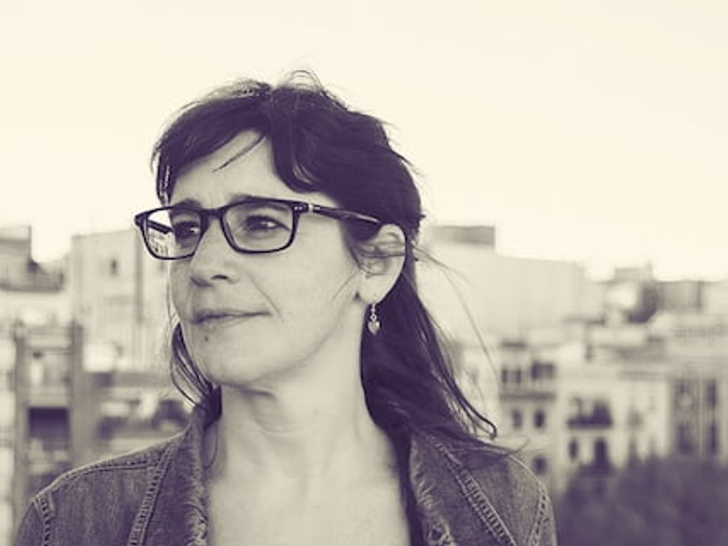 A black and white portrait of a woman with glasses looking away from the camera, standing in front of a cityscape.