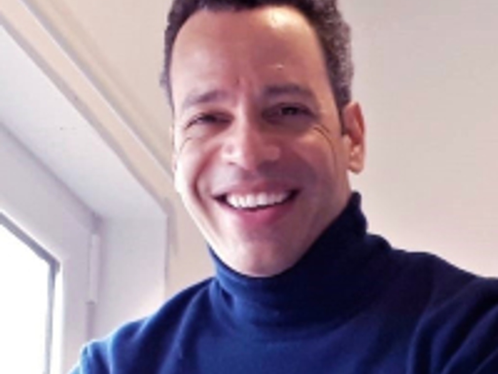 A smiling man wearing a blue turtleneck sweater in a bright indoor setting.