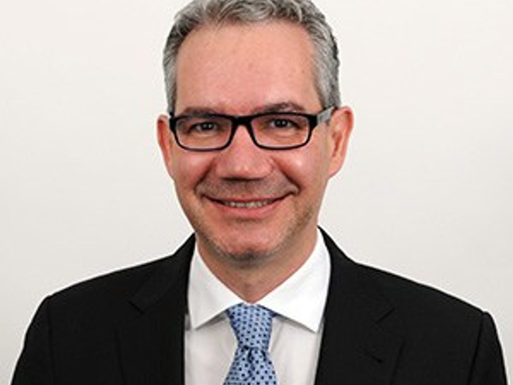 A professional portrait of a smiling man wearing glasses, a black suit, white shirt, and blue tie.