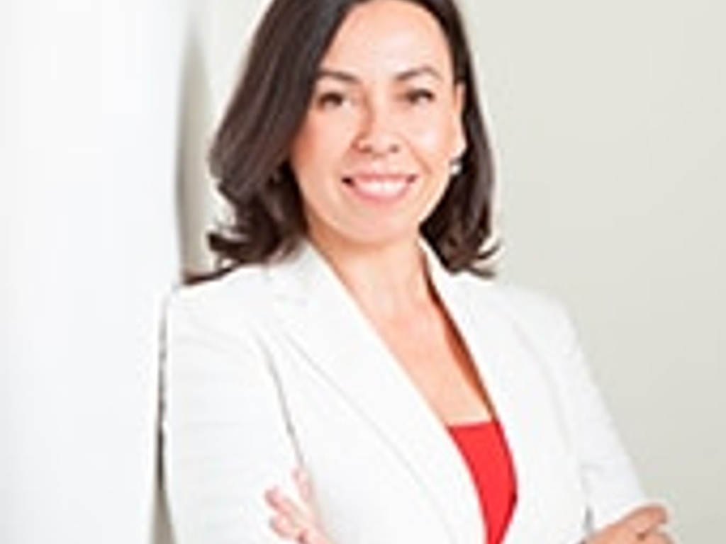 A professional woman in a white blazer and red top stands confidently with her arms crossed.