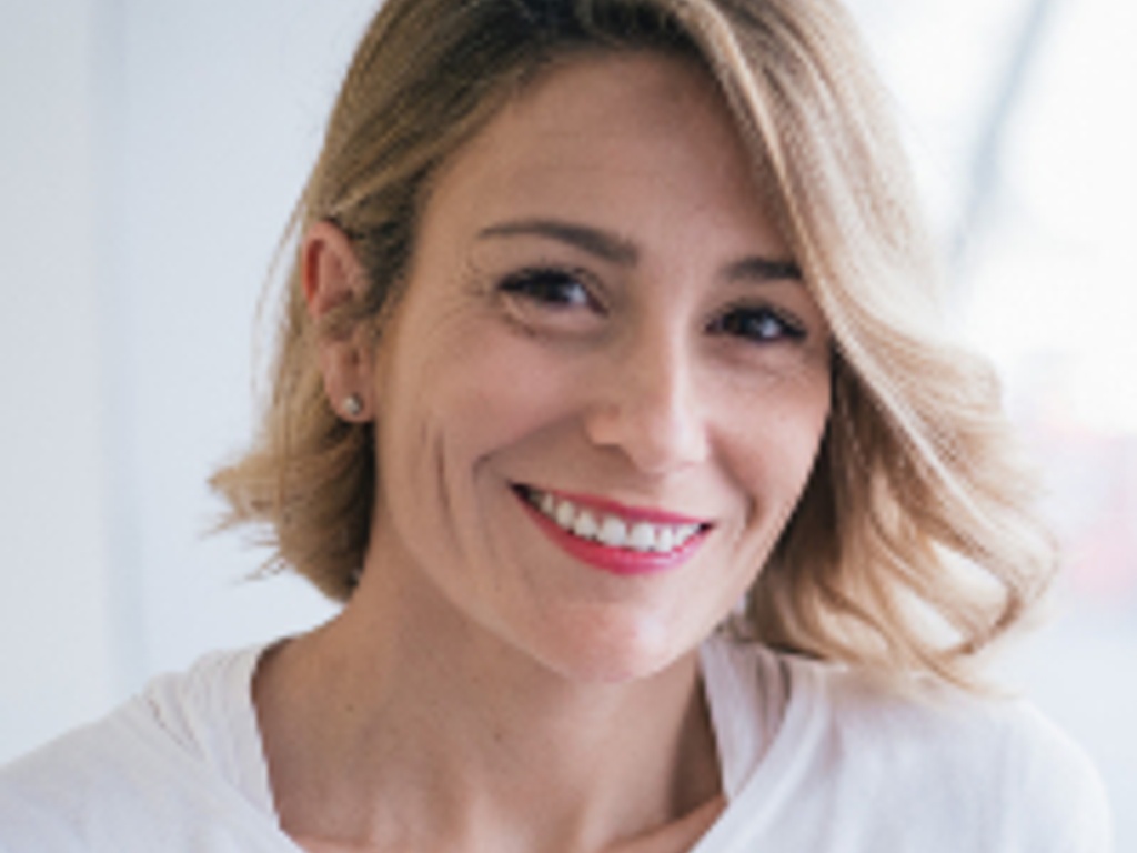 A portrait of a smiling woman with short blond hair, wearing a white t-shirt.