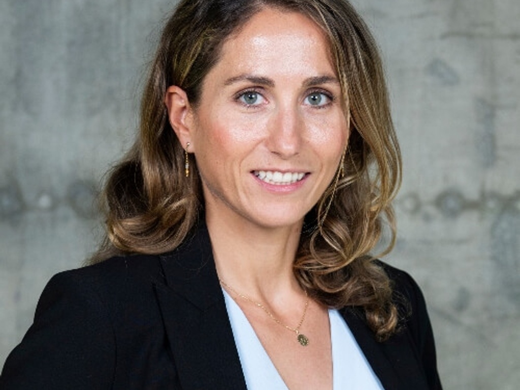 A professional portrait of a woman with long brown hair wearing a black blazer and white top, smiling against a gray background.