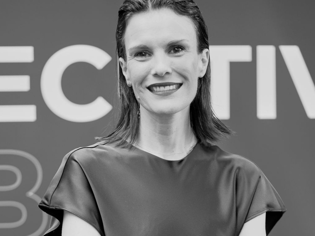 A black and white photo of a smiling woman with long hair, standing with her arms crossed in front of a sign that reads 'EFFECTIVE'.