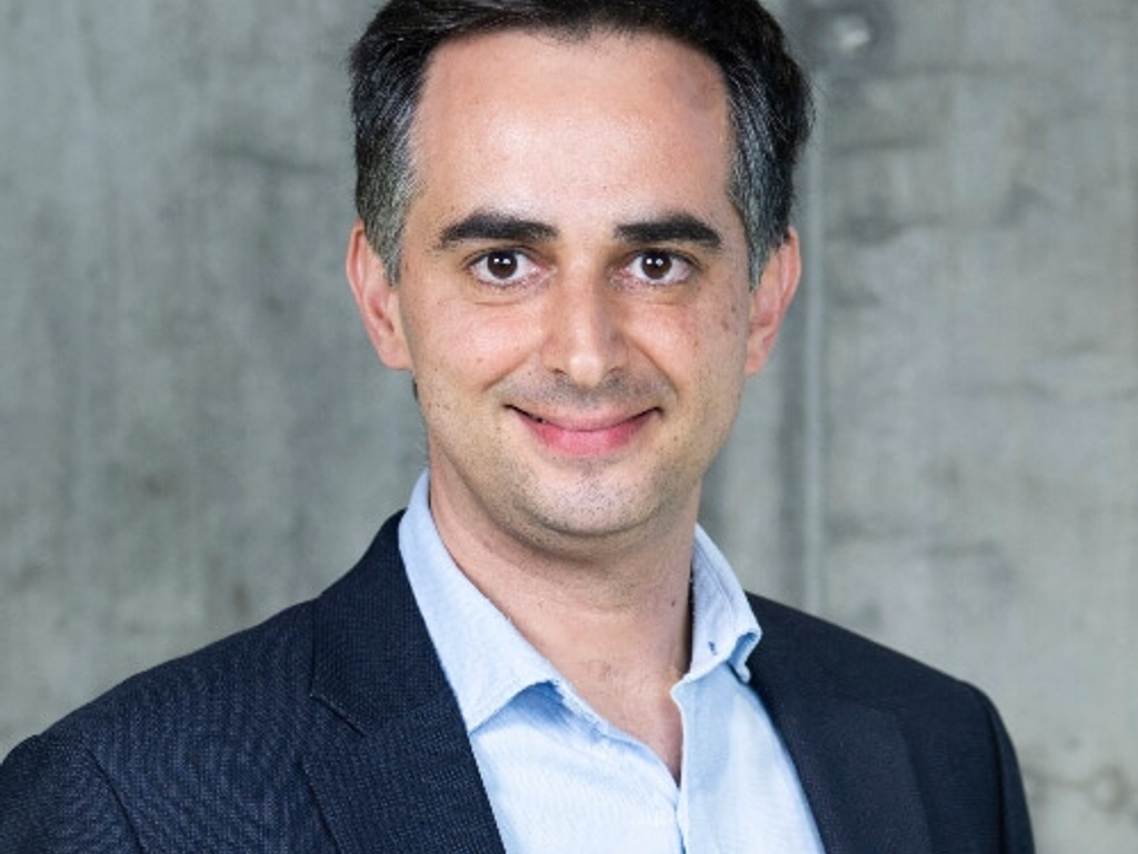 A professional portrait of a smiling man wearing a suit jacket and a blue shirt against a textured gray background.