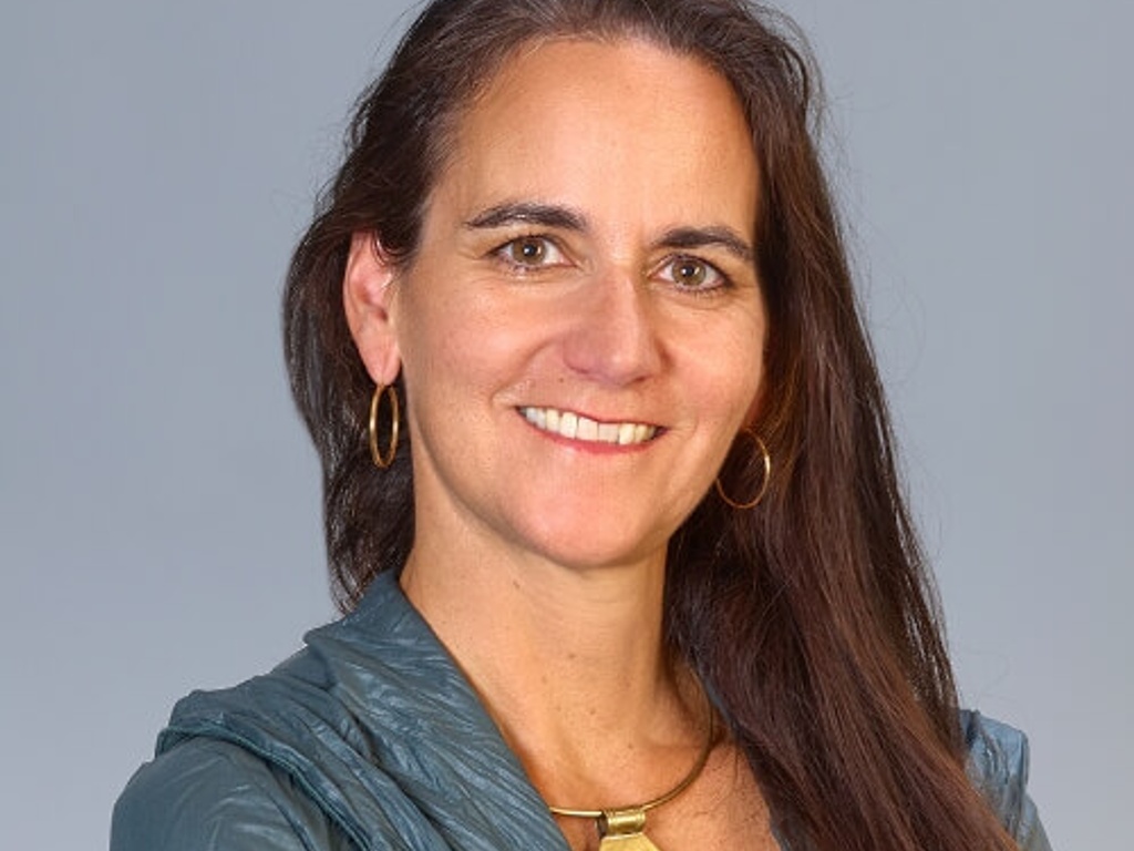 Portrait of a smiling woman with dark hair, wearing a teal blouse and a large golden necklace.