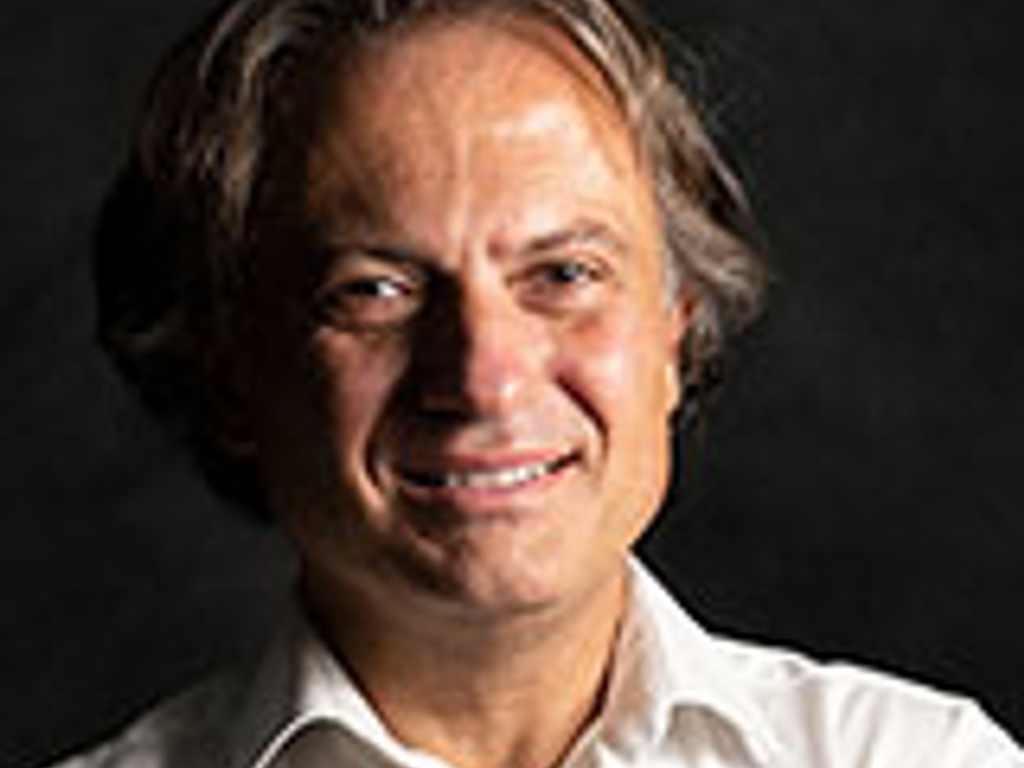 Portrait of a smiling middle-aged man with medium-length brown hair, wearing a white shirt against a dark background.