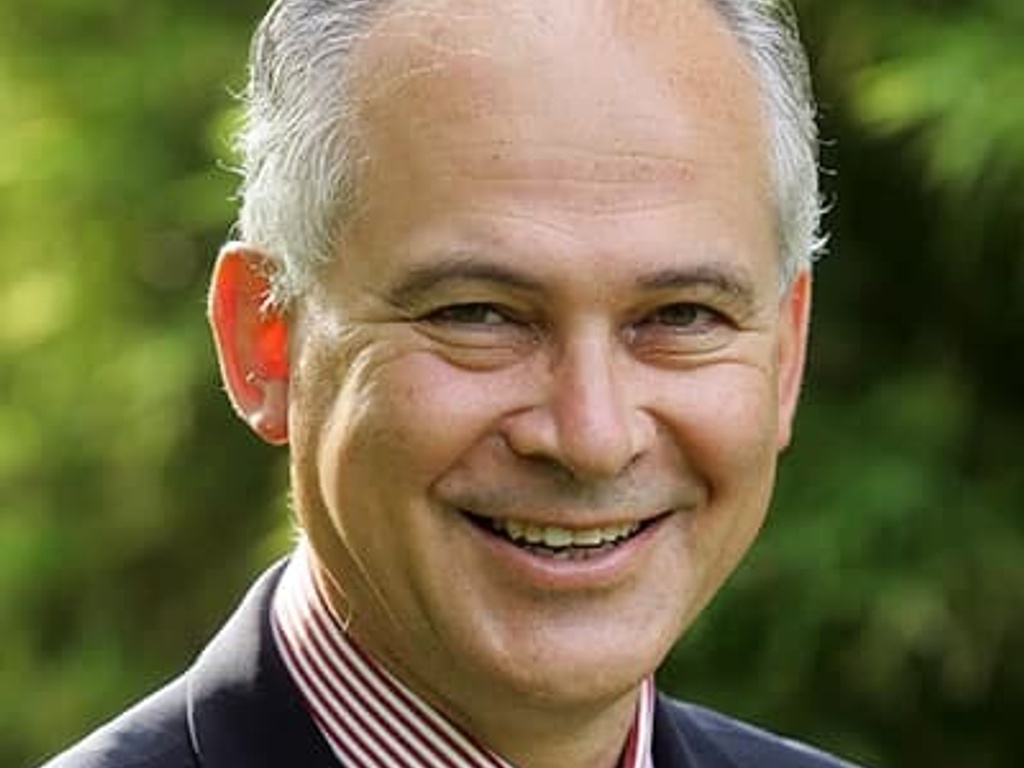 A portrait of a smiling middle-aged man wearing a suit and a red striped tie, set against a blurred green background.