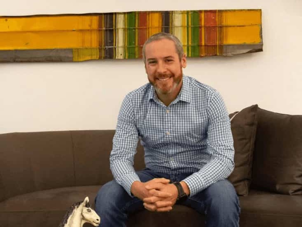 A smiling man in a blue checked shirt sitting on a dark sofa in a room with decorative items on the wall and on the table.