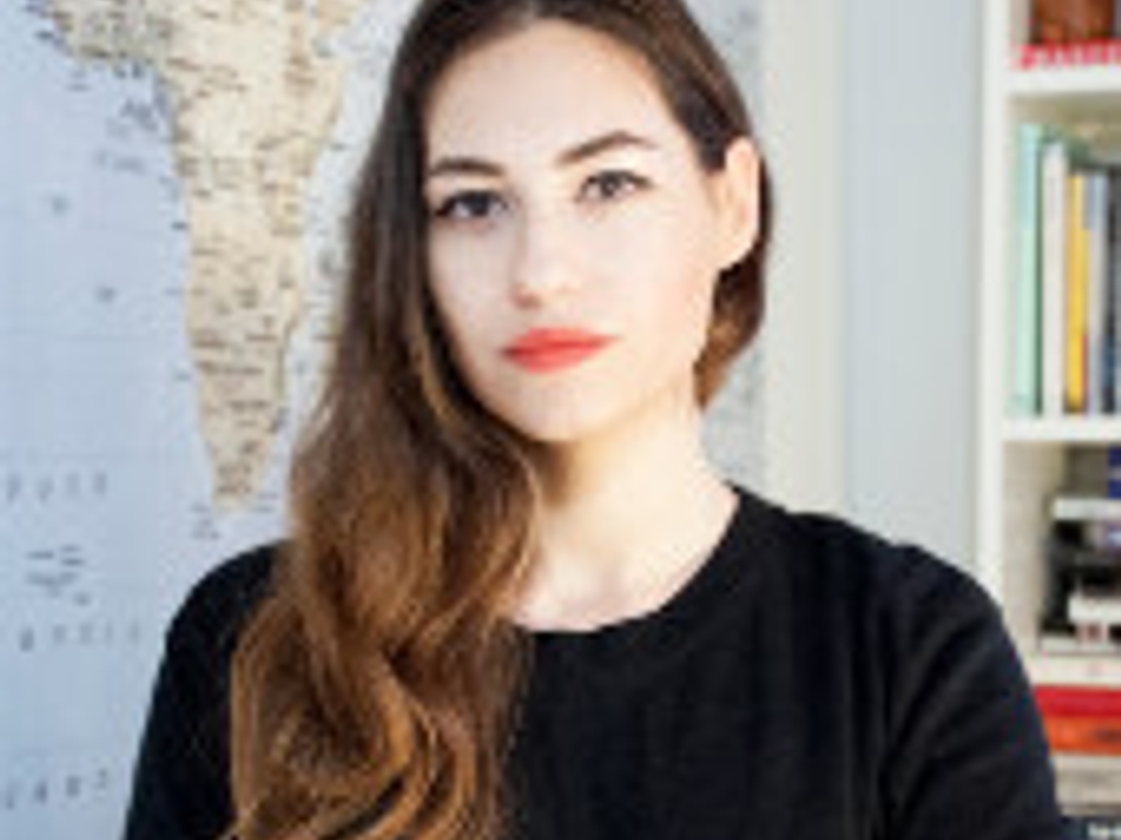 A woman with long hair wearing a black top stands in front of a bookshelf and a wall map.