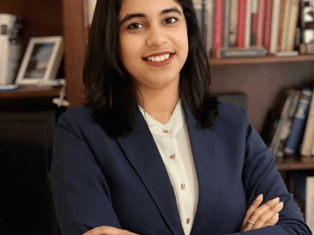 A smiling woman in a blue blazer stands confidently with arms crossed in front of a bookshelf.