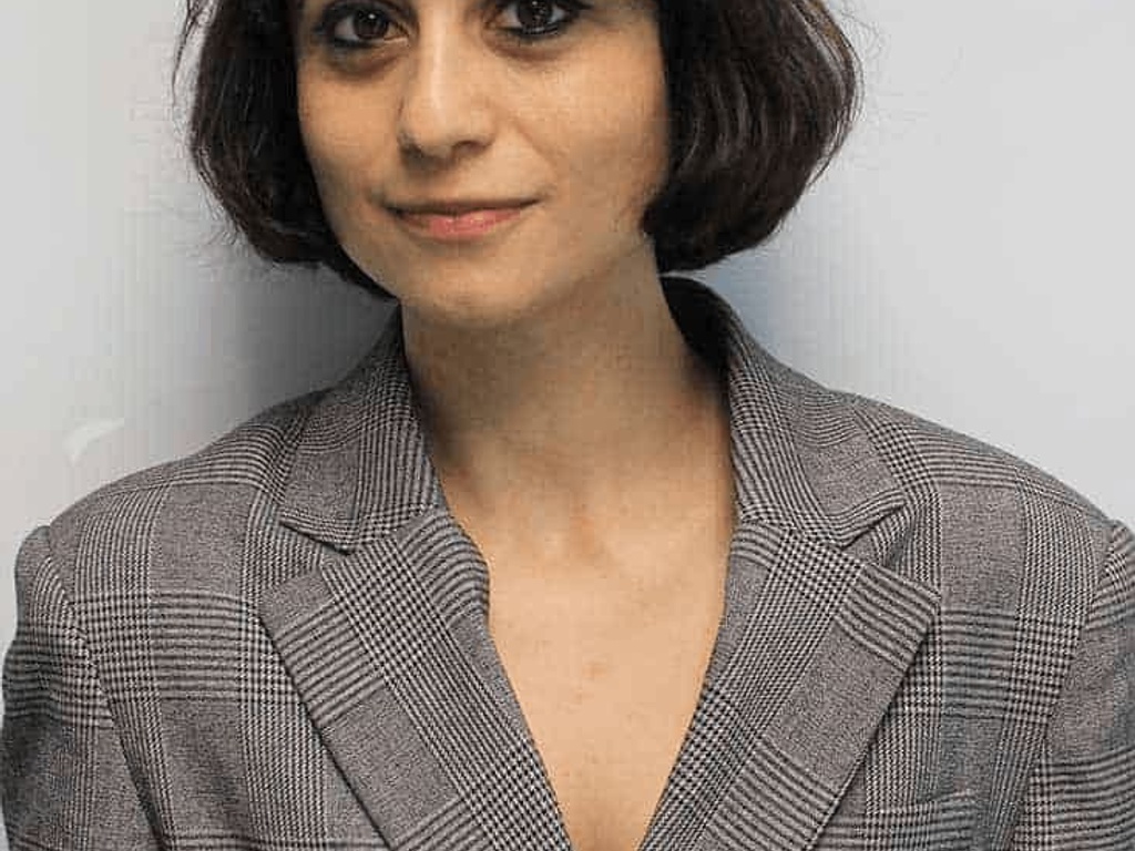 A professional portrait of a woman with short dark hair wearing a grey blazer, standing against a plain background.