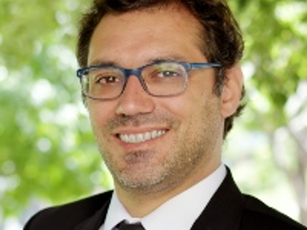 A portrait of a smiling man wearing glasses and a black suit standing outdoors with trees in the background.