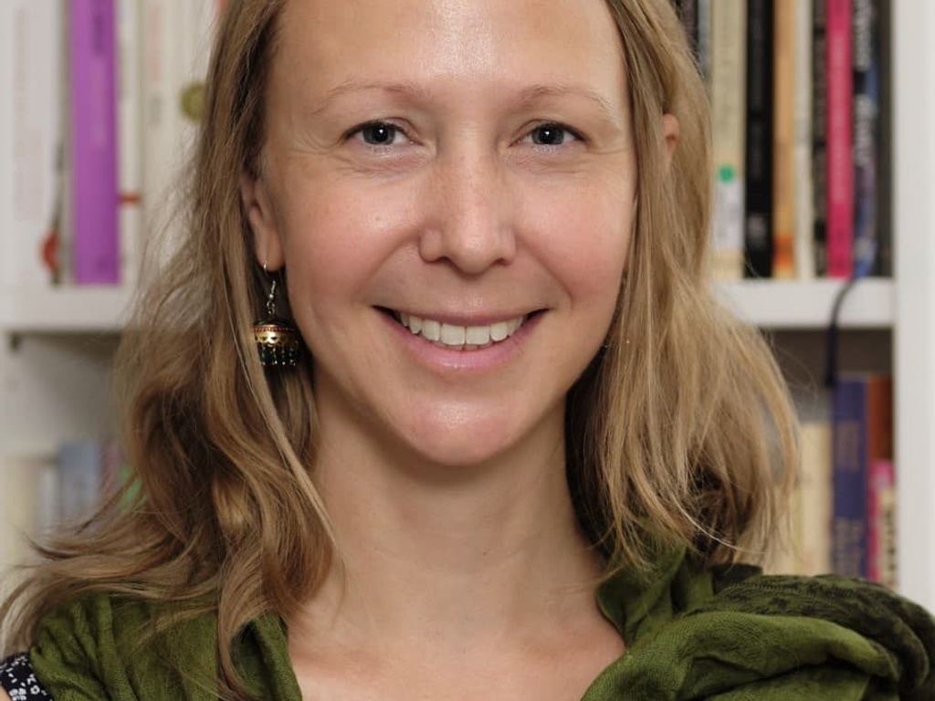 A woman with blond hair smiles gently, wearing a green scarf and standing in front of a bookshelf.