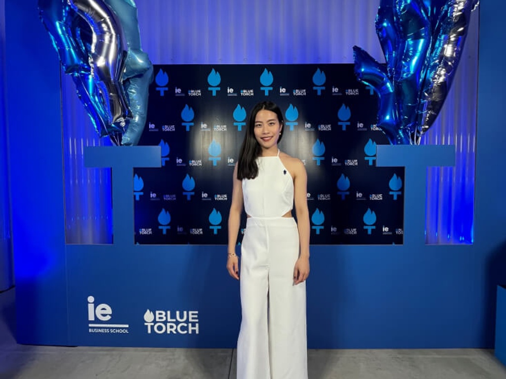 A woman in a white jumpsuit is standing in front of a promotional backdrop with blue balloons and logos.